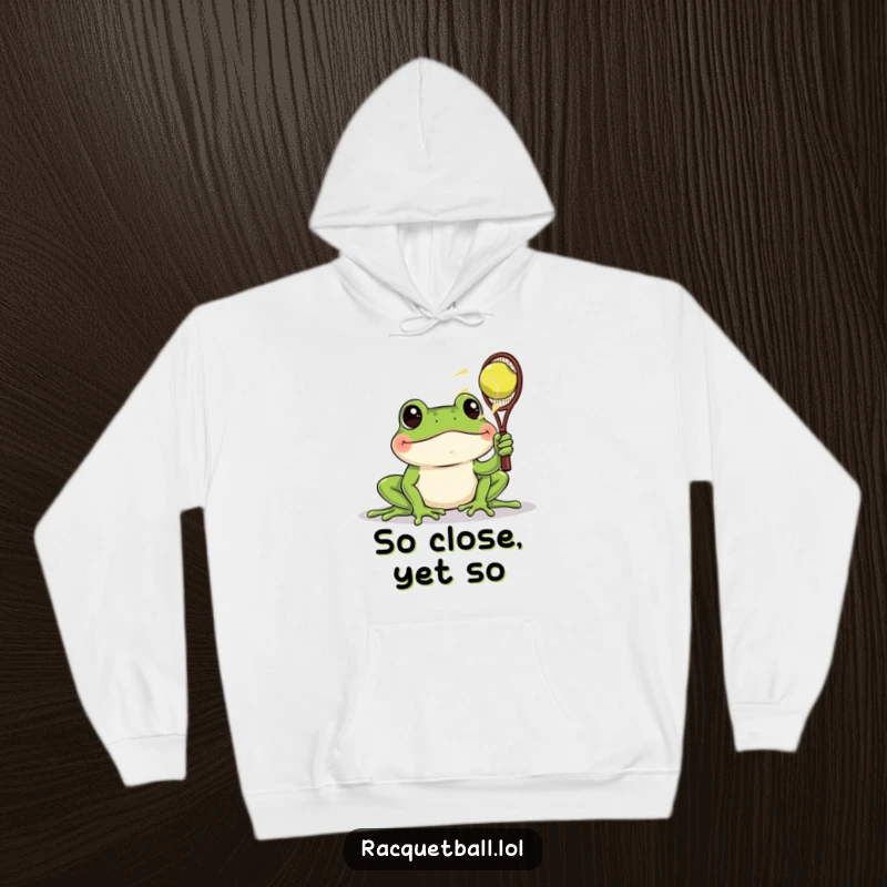 Funny hoodie with a frog intently watching a fast racquetball, embodying humorous observation and cozy comfort.